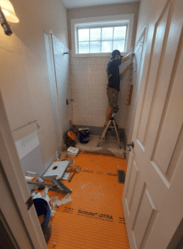 A person stands on a step ladder installing white tiles on a shower wall in a bathroom under renovation, with tools and materials on the floor.