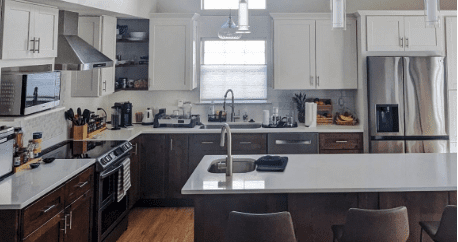 Modern kitchen with white upper cabinets, dark lower cabinets, stainless steel appliances, central island with sink, and various kitchen items on the counters.